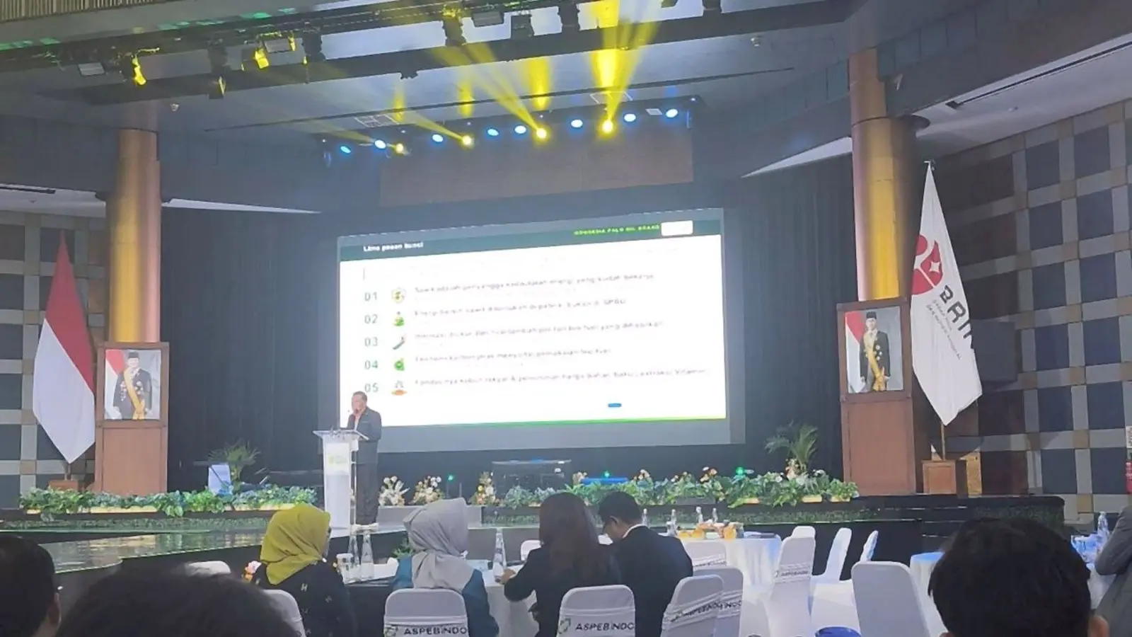 Tampak jauh seorang pembicara di podium dalam ruang konferensi.